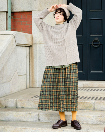 ウール本来の色味をいかした柄編みニットに、カーキのチェックスカートを合わせた、落ち着きのあるナチュラルコーデ。凹凸のあるシャーリング生地で仕立てたスカートは、歩くたびに軽やかに揺れ、やわらかな陰影を生み出します。自然体で過ごしたい日にぴったりの装い。