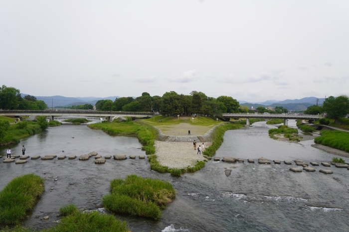 【画像は、賀茂大橋から望む賀茂川と高野川が合流する三角地。その北に広がる森は「下鴨神社」の境内の一部“糺の森”です。高野川（画像左）に架かるのが、「出町柳駅」前から続く“河合橋”、鴨川（画像中央から右）へ架かるのが“出町橋”。】